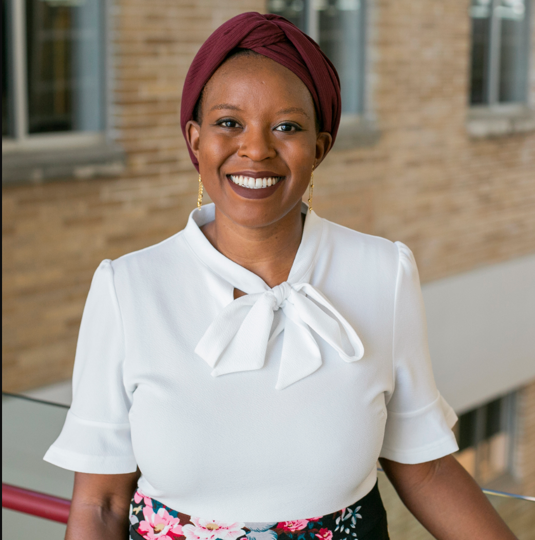 Photo of Prof. Nyamagaga Gondwe. She is standing in front of the Law School library interior. She is a Black woman wearing a white blouse and maroon head scarf.