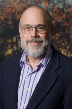 Photo of Prof. Peter Carstensen. He is a bald white man with a gray beard. He is wearing glasses and a black suit jacket.